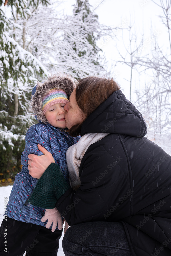 Mom with daughter in winter snowy forest. Mom kisses her daughter and warms her hands with her breath. Young girl with her daughter walking in the forest in winter.