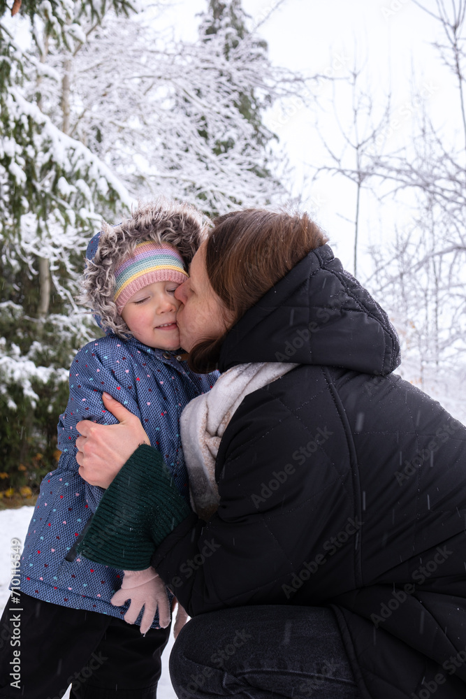 Mom with daughter in winter snowy forest. Mom kisses her daughter and warms her hands with her breath. Young girl with her daughter walking in the forest in winter. Mom with daughter in winter snowy forest. Mom kisses her daughter and warms her hands with her breath. Young girl with her daughter walking in the forest in winter.