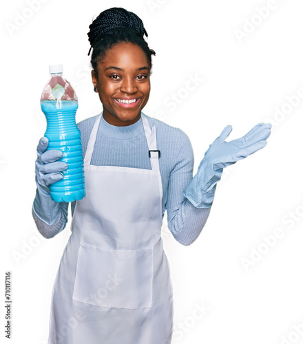 Young african american woman wearing apron holding detergent bottle celebrating victory with happy smile and winner expression with raised hands