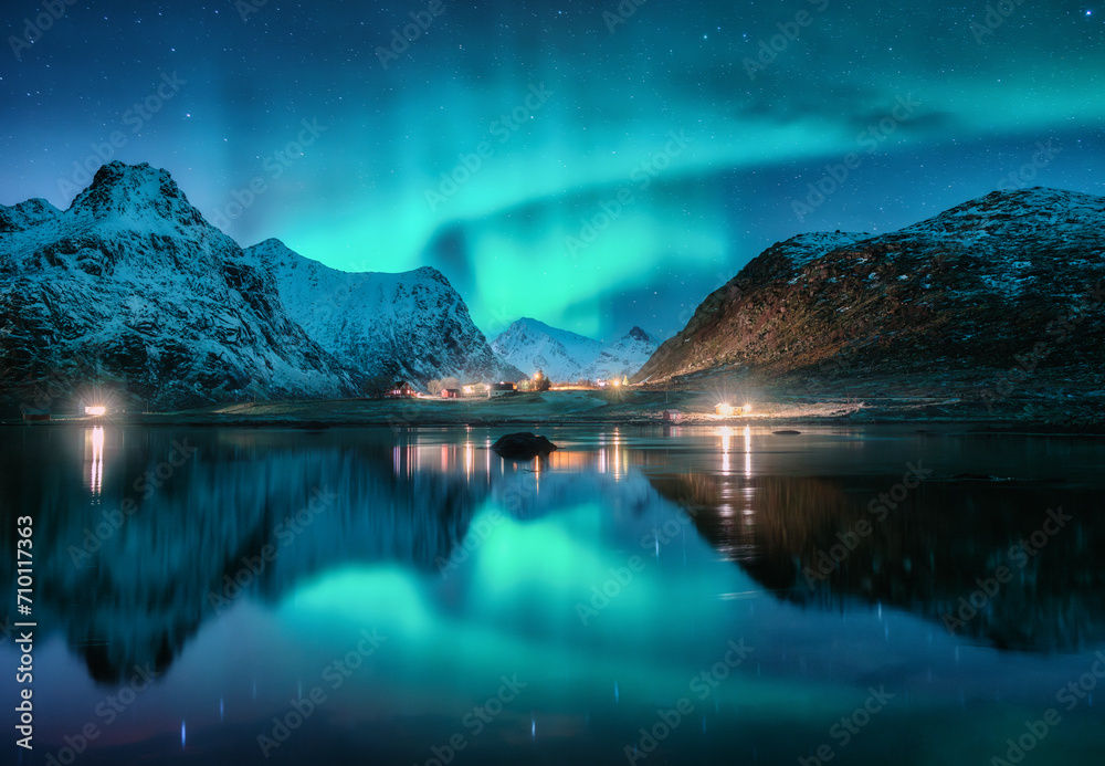 Aurora borealis, snowy mountains, sea, fjord, reflection in water, street lights at starry winter night. Lofoten, Norway. Northern lights. Landscape with polar lights, snowy rocks, sky with stars
