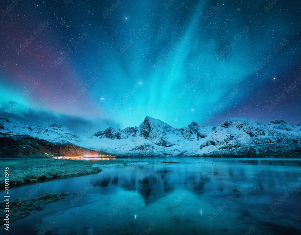 Northern lights over the snowy mountains, frozen sea, reflection in water at winter night in Lofoten, Norway. Aurora borealis and snowy rocks. Landscape with polar lights, starry sky and fjord. Nature