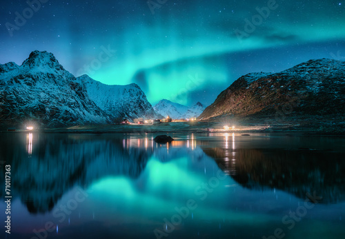 Aurora borealis, snowy mountains, sea, fjord, reflection in water, street lights at starry winter night. Lofoten, Norway. Northern lights. Landscape with polar lights, snowy rocks, sky with stars