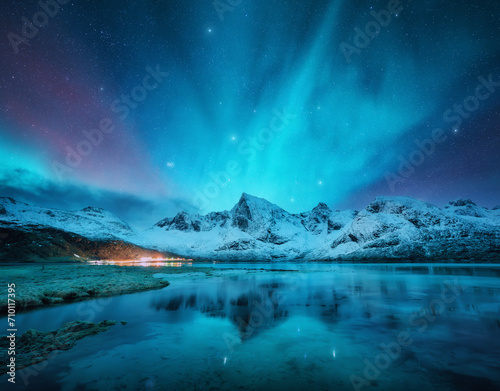Northern lights over the snowy mountains, frozen sea, reflection in water at winter night in Lofoten, Norway. Aurora borealis and snowy rocks. Landscape with polar lights, starry sky and fjord. Nature photo