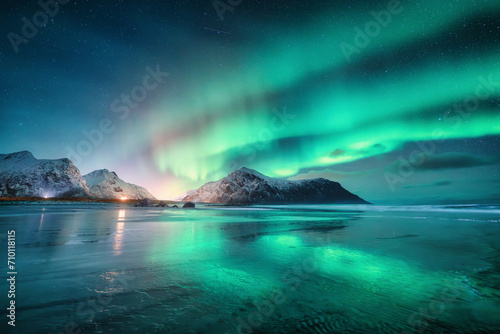 Northern Lights and sandy beach at starry winter night. Lofoten islands, Norway. Beautiful Aurora borealis. Sky with polar lights. Landscape with aurora, sea, sky reflection in water, snowy mountains © den-belitsky