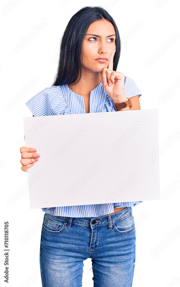 Young beautiful latin girl holding blank empty banner serious face thinking about question with hand on chin, thoughtful about confusing idea Young beautiful latin girl holding blank empty banner serious face thinking about question with hand on chin, thoughtful about confusing idea