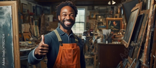 An African American painter in his studio, holding an empty frame, smiles happily and gives a thumbs up, expressing excellence and approval.