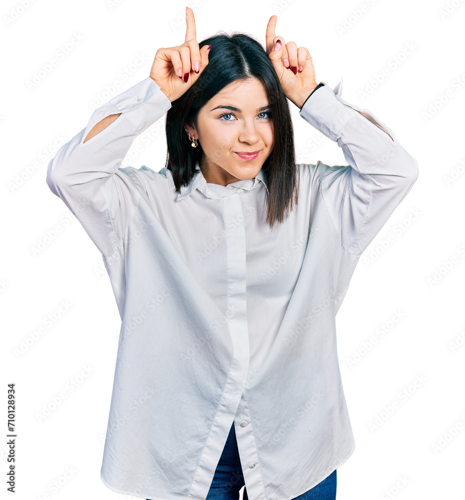Young brunette woman with blue eyes wearing oversize white shirt doing funny gesture with finger over head as bull horns Young brunette woman with blue eyes wearing oversize white shirt doing funny gesture with finger over head as bull horns