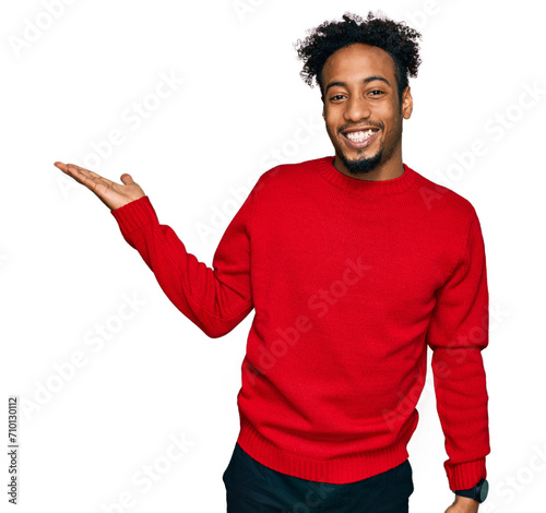 Young african american man with beard wearing casual winter sweater smiling cheerful presenting and pointing with palm of hand looking at the camera.