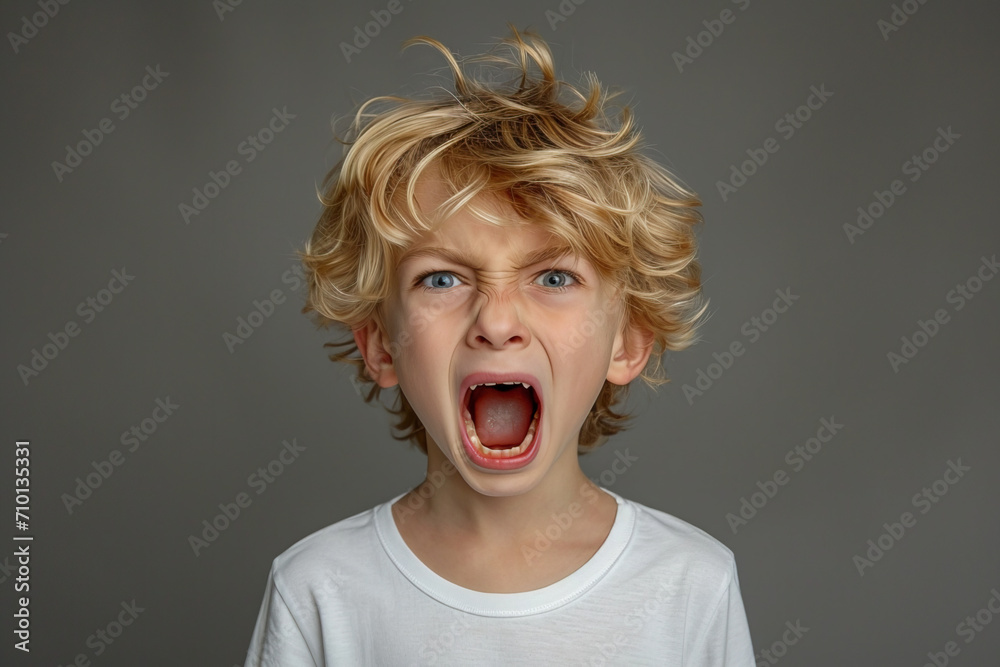 A portrait of a European boy with blond hair who is screaming with his mouth wide open, with an unhappy and aggressive expression. He stands on a gray studio background