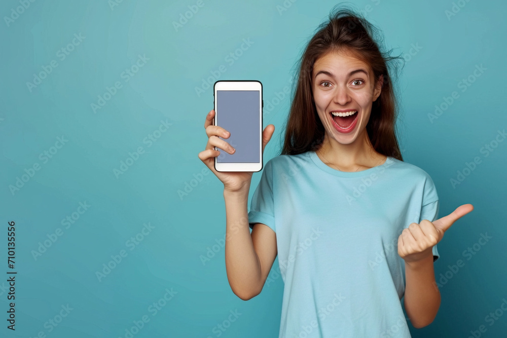 An excited young brunette in a blue T-shirt on a blue background is holding a smartphone with a blank screen and pointing at it. Advertising concept