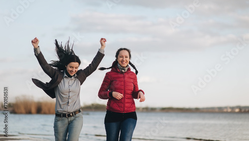 Two girlfriends in blue jeans, a brown and red jackets and a gray shirt run along the river bank in the evening of sunset and laugh with joy. The concept of a spring walk outdoors.
