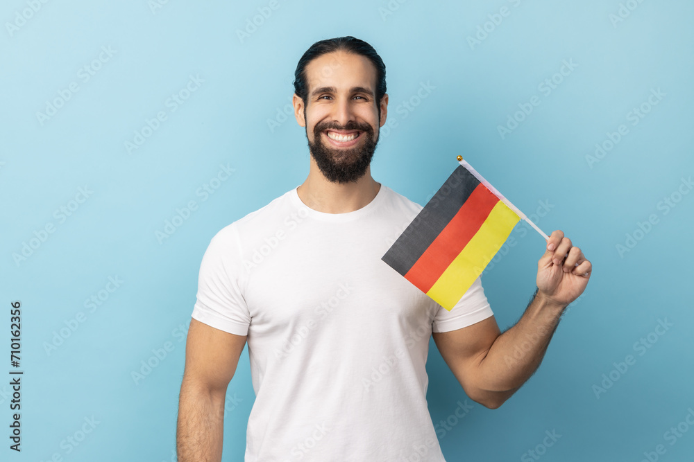 Portrait of positive smiling man with beard wearing white T-shirt holding Germany flag, celebrating Day of Germany - 3th October, looking at camera. Indoor studio shot isolated on blue background. Portrait of positive smiling man with beard wearing white T-shirt holding Germany flag, celebrating Day of Germany - 3th October, looking at camera. Indoor studio shot isolated on blue background.