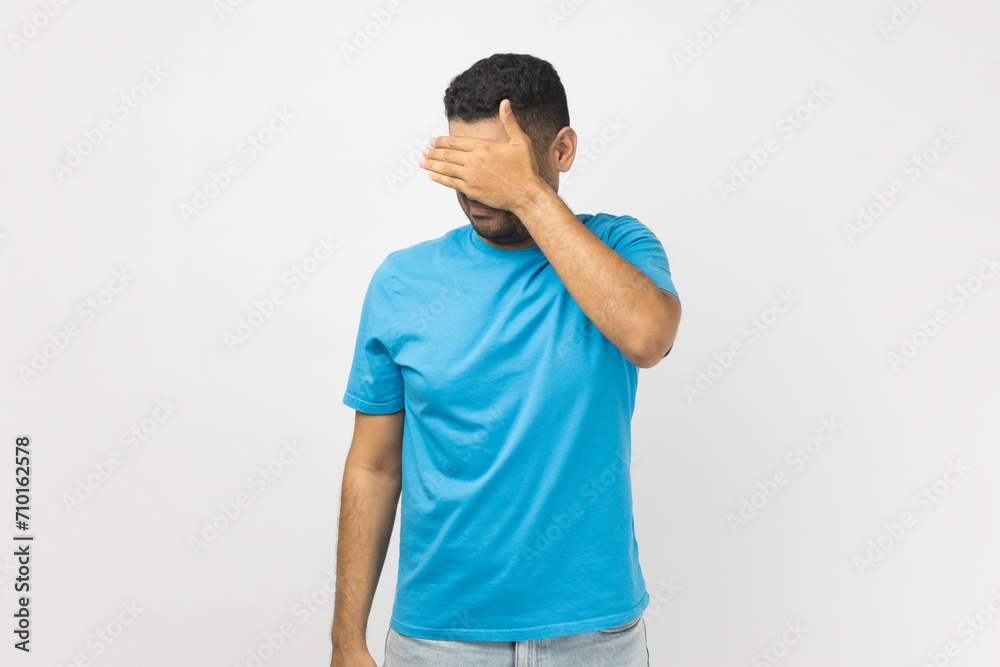 Portrait of shy young adult unshaven man wearing blue T- shirt standing covering his face with hand, hiding his face, doesn't want to see. Indoor studio shot isolated on gray background. Portrait of shy young adult unshaven man wearing blue T- shirt standing covering his face with hand, hiding his face, doesn't want to see. Indoor studio shot isolated on gray background.