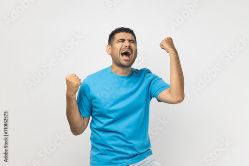 Portrait of unshaven man wearing blue T- shirt standing clenches fists with positive expression, rejoices wonderful news, smiles happily. Indoor studio shot isolated on gray background. photo