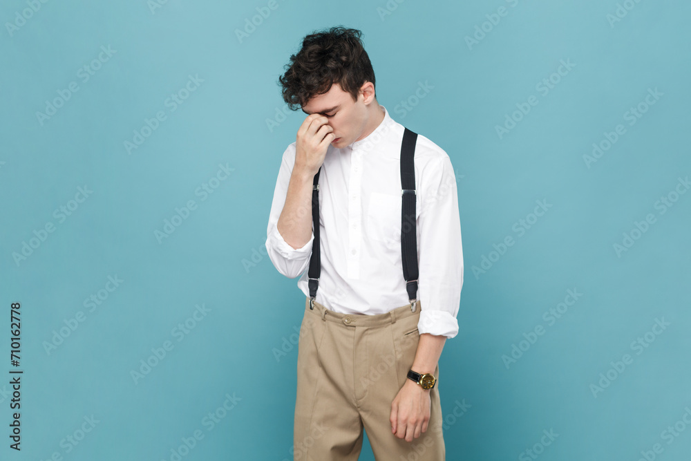 Portrait of sad upset disappointed man wearing white shirt and suspender standing and crying having serious problems, being in bad mood. Indoor studio shot isolated on blue background.