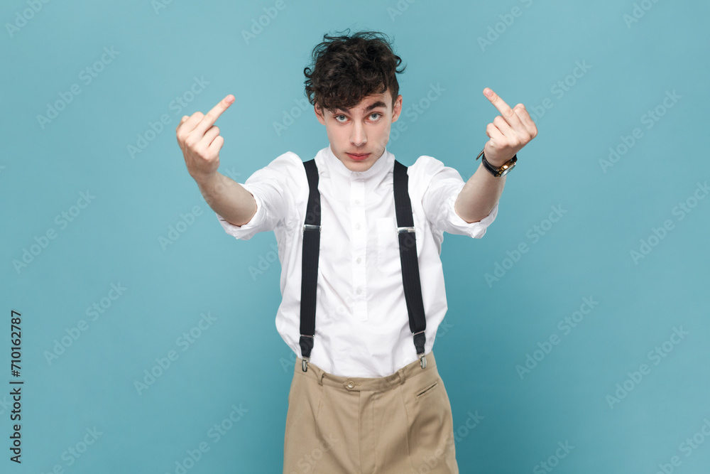 Portrait of aggressive man wearing white shirt and suspender showing middle finger, rude gesture, face full of hatred and resentment. Indoor studio shot isolated on blue background.
