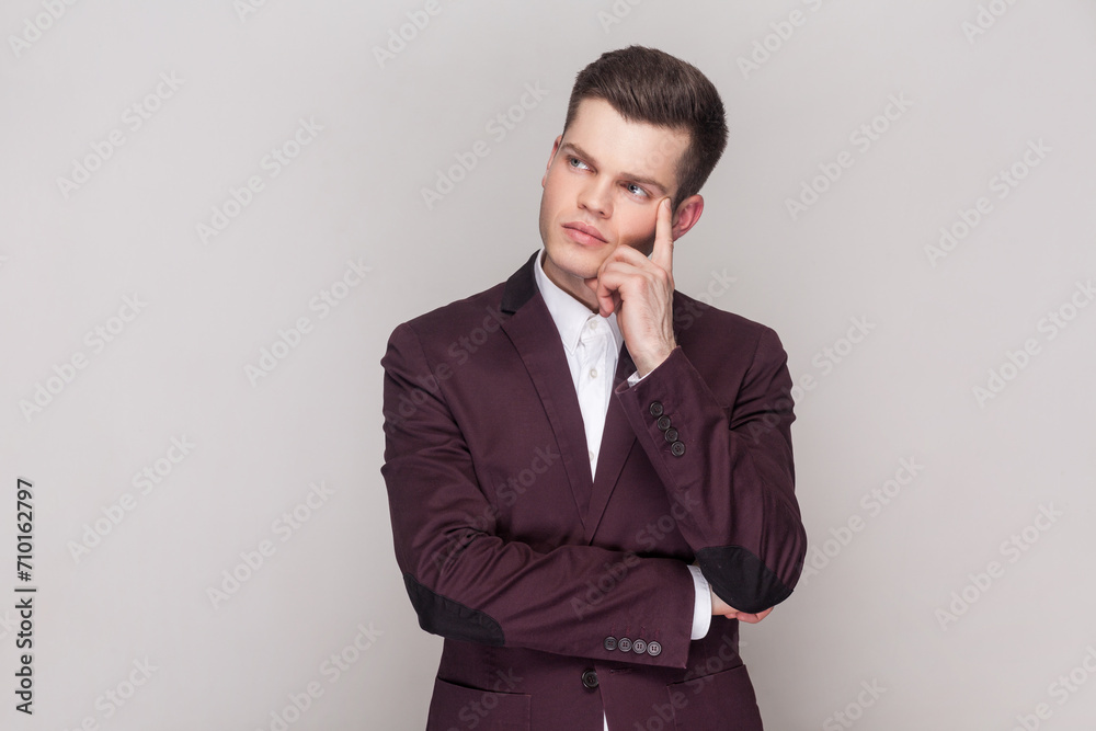 Pensive handsome young man keeps finger on temple, being deep in thoughts, has serious face, remembers something, wearing violet suit and white shirt. Indoor studio shot isolated on grey background. Pensive handsome young man keeps finger on temple, being deep in thoughts, has serious face, remembers something, wearing violet suit and white shirt. Indoor studio shot isolated on grey background.