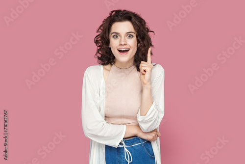 Portrait of clever smart happy woman with curly hair wearing casual style outfit raises her index finger up, having good idea. Indoor studio shot isolated on pink background. photo