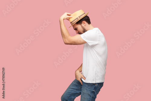 Portrait of handsome bearded hipster man in white shirt and casual hat standing in michael jackson dancing pose, being in good happy mood. Indoor studio shot isolated on pink background.