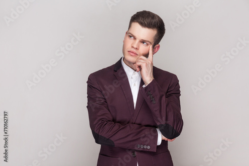 Pensive handsome young man keeps finger on temple, being deep in thoughts, has serious face, remembers something, wearing violet suit and white shirt. Indoor studio shot isolated on grey background.