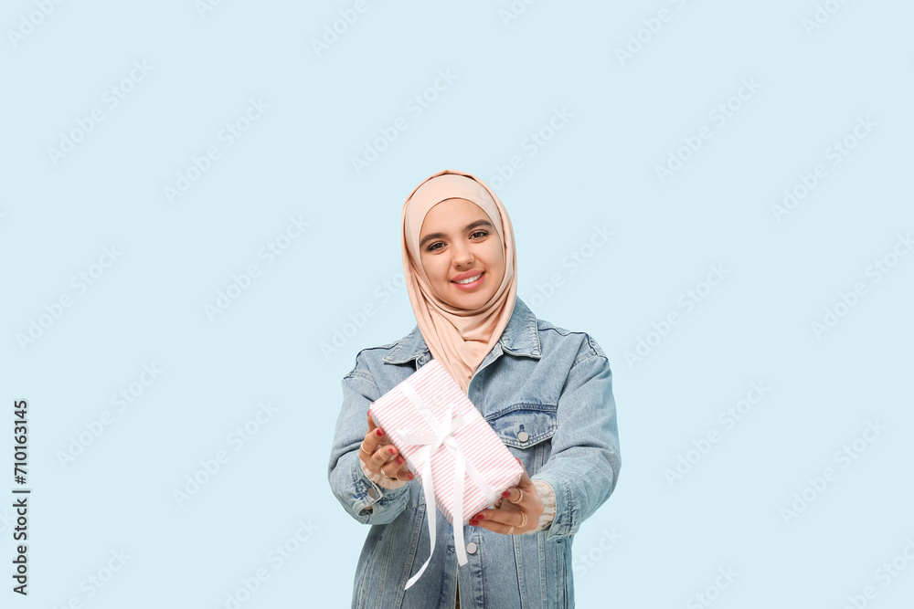 Happy young Muslim woman in hijab with gift box on blue background