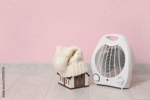 Decorative house with hat and electric fan heater near pink wall. Winter heating concept photo