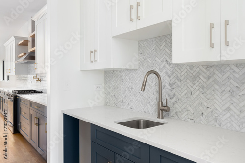 A scullery or butler's pantry detail with a bronze faucet and hardware, blue and white cabinets, and marble herringbone tile backsplash and countertop. photo