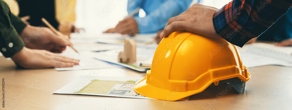 Professional architect team and engineer discuss about architectural project on meeting table with safety helmet, wooden block and architectural document scatter around. Closeup. Delineation. - obrazy, fototapety, plakaty 