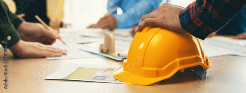 Fototapeta Professional architect team and engineer discuss about architectural project on meeting table with safety helmet, wooden block and architectural document scatter around. Closeup. Delineation.