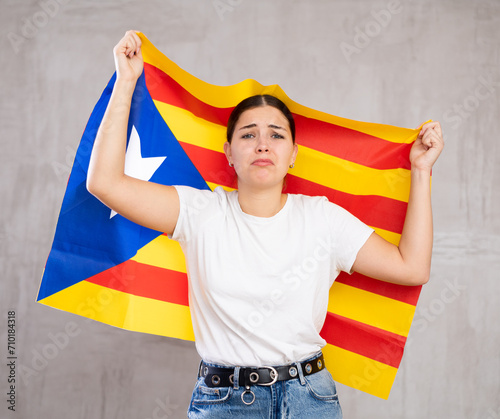 Sad young woman with Catalonia flag in hands posing sorrowfully against light unicoloured background
