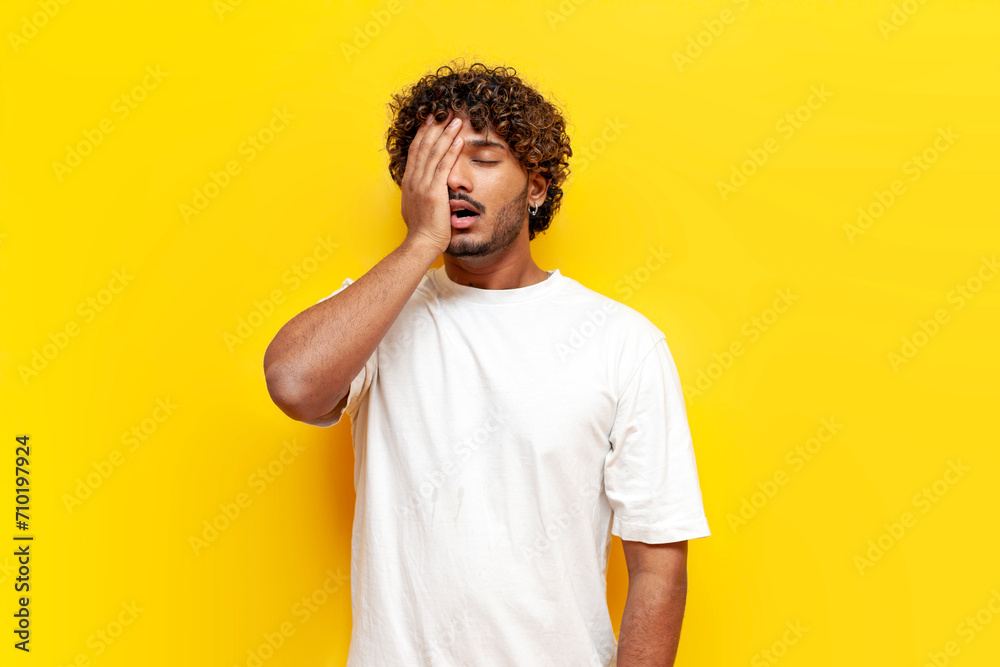 young curly indian man showing facepalm and failure on yellow isolated background, sad guy lost and covering his face with his hand from making a mistake young curly indian man showing facepalm and failure on yellow isolated background, sad guy lost and covering his face with his hand from making a mistake