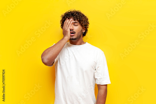 young curly indian man showing facepalm and failure on yellow isolated background, sad guy lost and covering his face with his hand from making a mistake