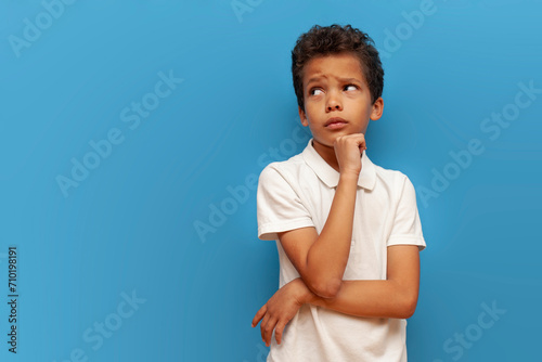 african american sad teenager boy in white polo thinks and plans on blue isolated background, unhappy pensive child dreams and imagines