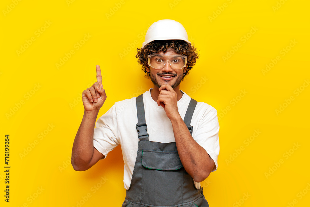 young indian male builder in a hard hat and overalls thinks and points with his finger up on a yellow isolated background, a foreman in a uniform and safety glasses chooses an idea young indian male builder in a hard hat and overalls thinks and points with his finger up on a yellow isolated background, a foreman in a uniform and safety glasses chooses an idea