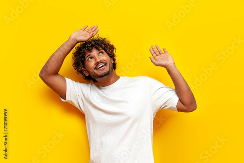 young Indian man fearfully raises his hands up and protects himself from falling from above on a yellow isolated background, the guy carries and catches with his hands up
