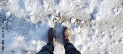 Male legs in jeans and black winter boots seen from above, standing on a snowy road on a sunny day.