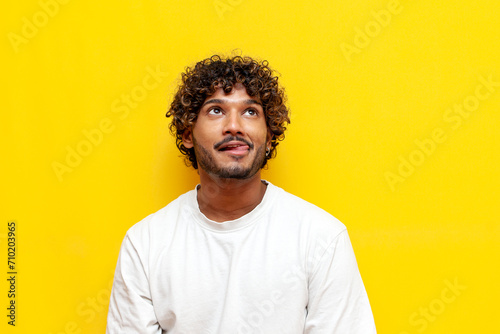 young curly indian man licking his lips and imagining on a yellow isolated background, a curious guy is dreaming and thinking looking up and showing his tongue photo