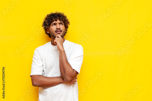 young pensive indian man in white t-shirt planning and thinking over yellow isolated background, curly puzzled guy imagining and dreaming. High quality photo photo