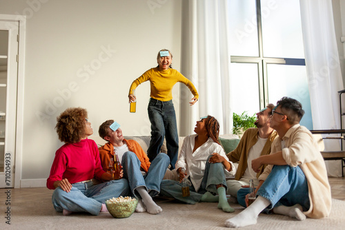 multiracial group of young people sitting at home with beer and popcorn and playing charades and having fun with friends, Asian girl showing and pretending to joke