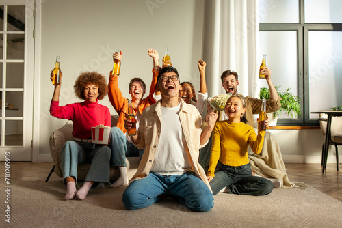 multiracial group of young friends sitting on the couch at home drinking beer and watching TV and celebrating good luck, students drinking alcohol rejoicing in victory and cheering and screaming