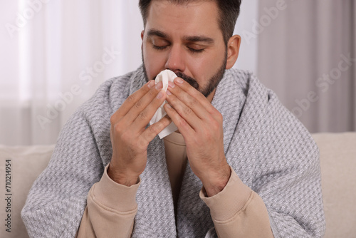 Sick man wrapped in blanket with tissue blowing nose on sofa indoors. Cold symptoms