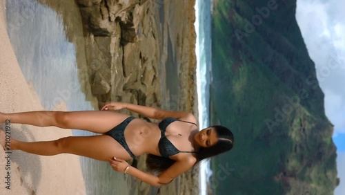 A beautiful woman in a sexy bikini poses gracefully near the rocks at the edge of the sea. Vertical video. photo