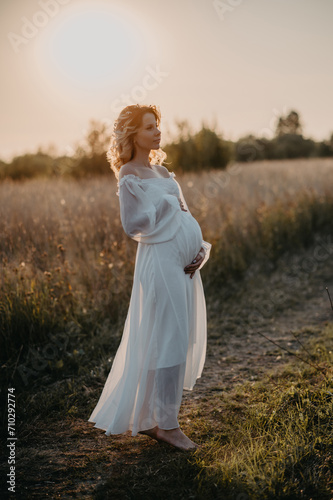 A young expectant mother in the third trimester of pregnancy in a white dress hugs her stomach against the backdrop of a natural landscape. The concept of future motherhood.