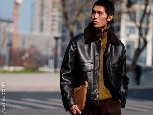 Portrait of handsome Chinese young man with black leather jacket posing with modern city street background in sunny winter day, male fashion, cool Asian young man.