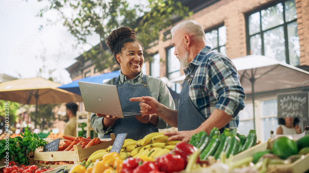 Partners Working at a Farmers Market. Middle Aged Couple Selling Ecological Fruits and Vegetables From a Food Stand. Street Vendors Using Laptop Computer and Discussing Steps for Business Development Partners Working at a Farmers Market. Middle Aged Couple Selling Ecological Fruits and Vegetables From a Food Stand. Street Vendors Using Laptop Computer and Discussing Steps for Business Development