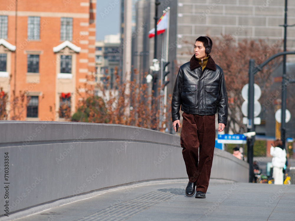 Φωτογραφία Portrait of handsome Chinese young man with black leather jacket wearing headset holding mobile phone posing with modern city street background in sunny winter day, male fashion, cool Asian young man