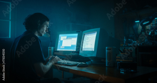 Caucasian Male Programmer Using Old Desktop Computer With Two Monitors In Retro Garage Late At Night. Evil Hacker Searching For Software Vulnerabilities, Coding DDOS Attack, Trojan Horse Program. © Gorodenkoff