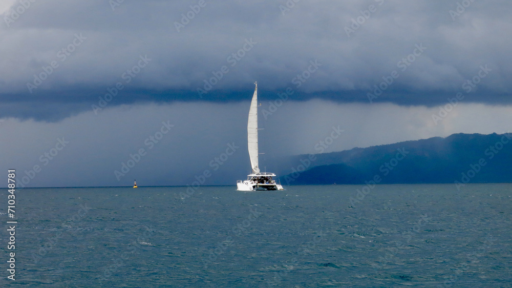 Naklejka premium A catamaran with a raised sail sails across the blue sea towards the rain. A sailing catamaran moves across the sea among tropical islands towards a storm on a cloudy day.