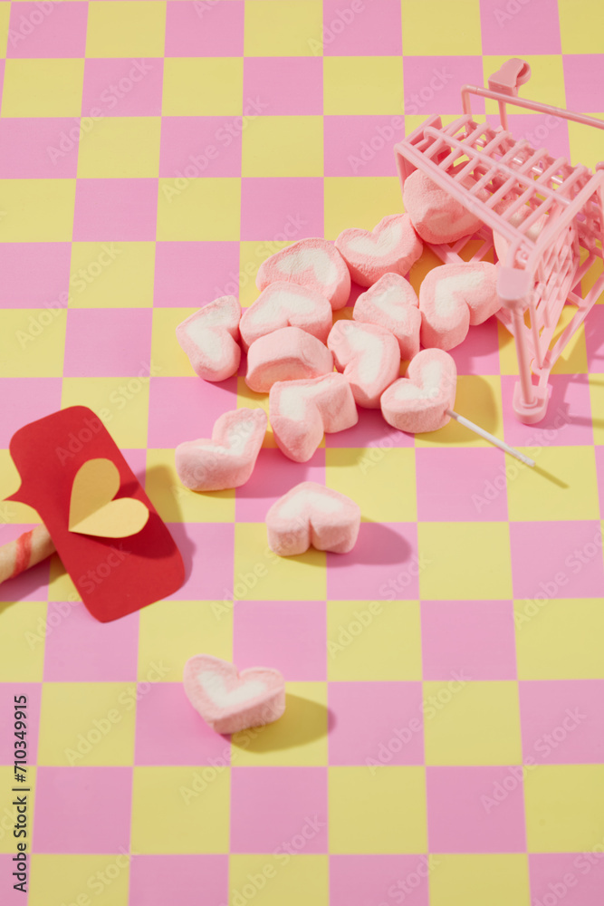 Pink mini trolley with sweet marshmallow in heart shape decorated on pink and yellow checkered background. Cute decorations for valentine's day. Copy space Pink mini trolley with sweet marshmallow in heart shape decorated on pink and yellow checkered background. Cute decorations for valentine's day. Copy space