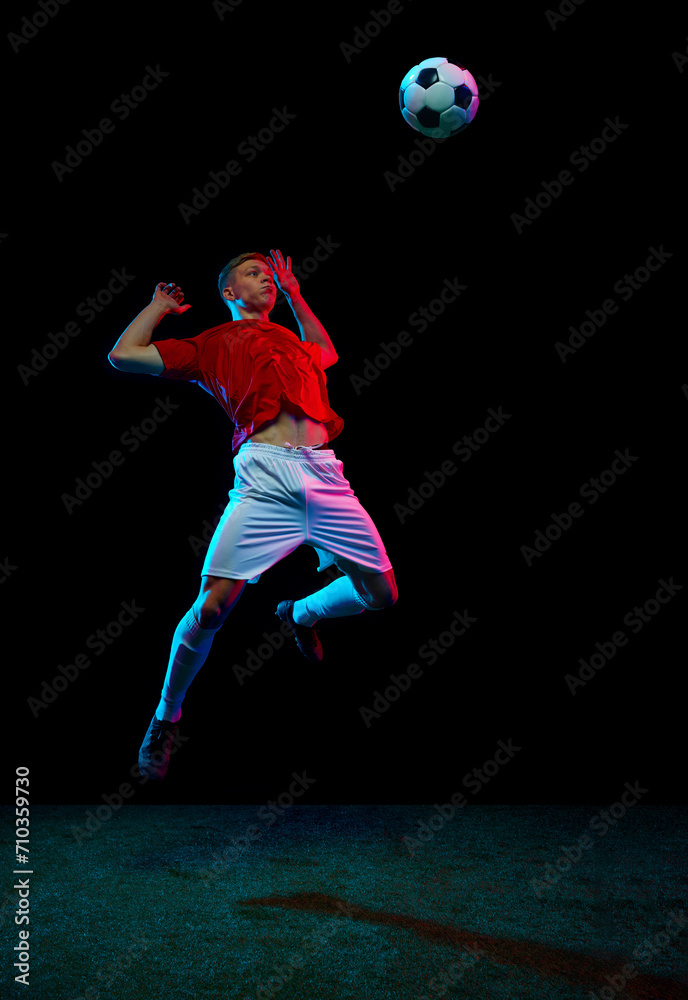 Airborne Mastery. Portrait of soccer as professional player executes flawless mid-air kick, sending ball soaring against black background in neon light. Concept of sport games, hobby, energy. movement Airborne Mastery. Portrait of soccer as professional player executes flawless mid-air kick, sending ball soaring against black background in neon light. Concept of sport games, hobby, energy. movement